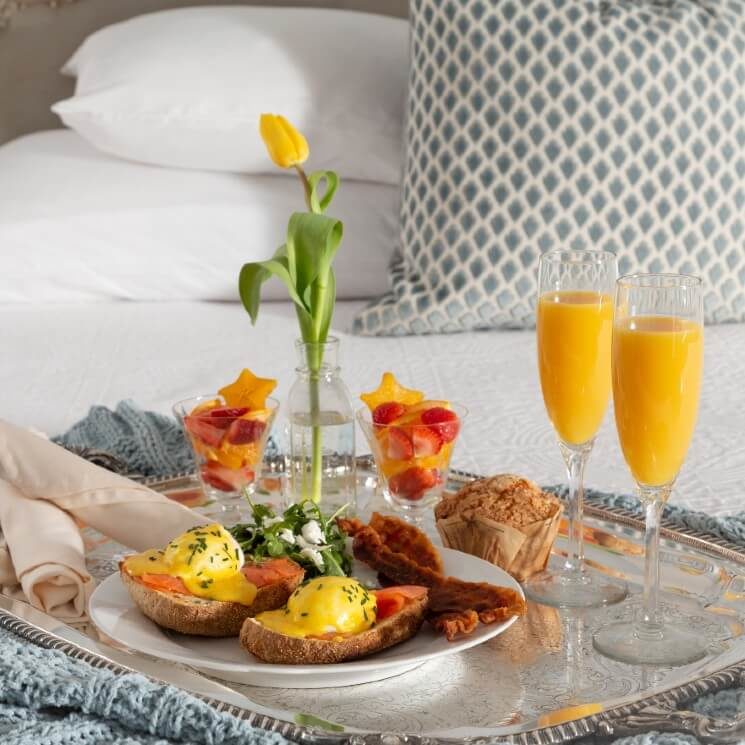 Silver tray on a bed holding a gourmet breakfast plate, flutes of orange juice and a flower