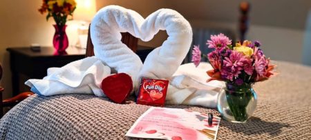A white towel in the shape of a heart on a bed with vase of flowers, candy and a letter