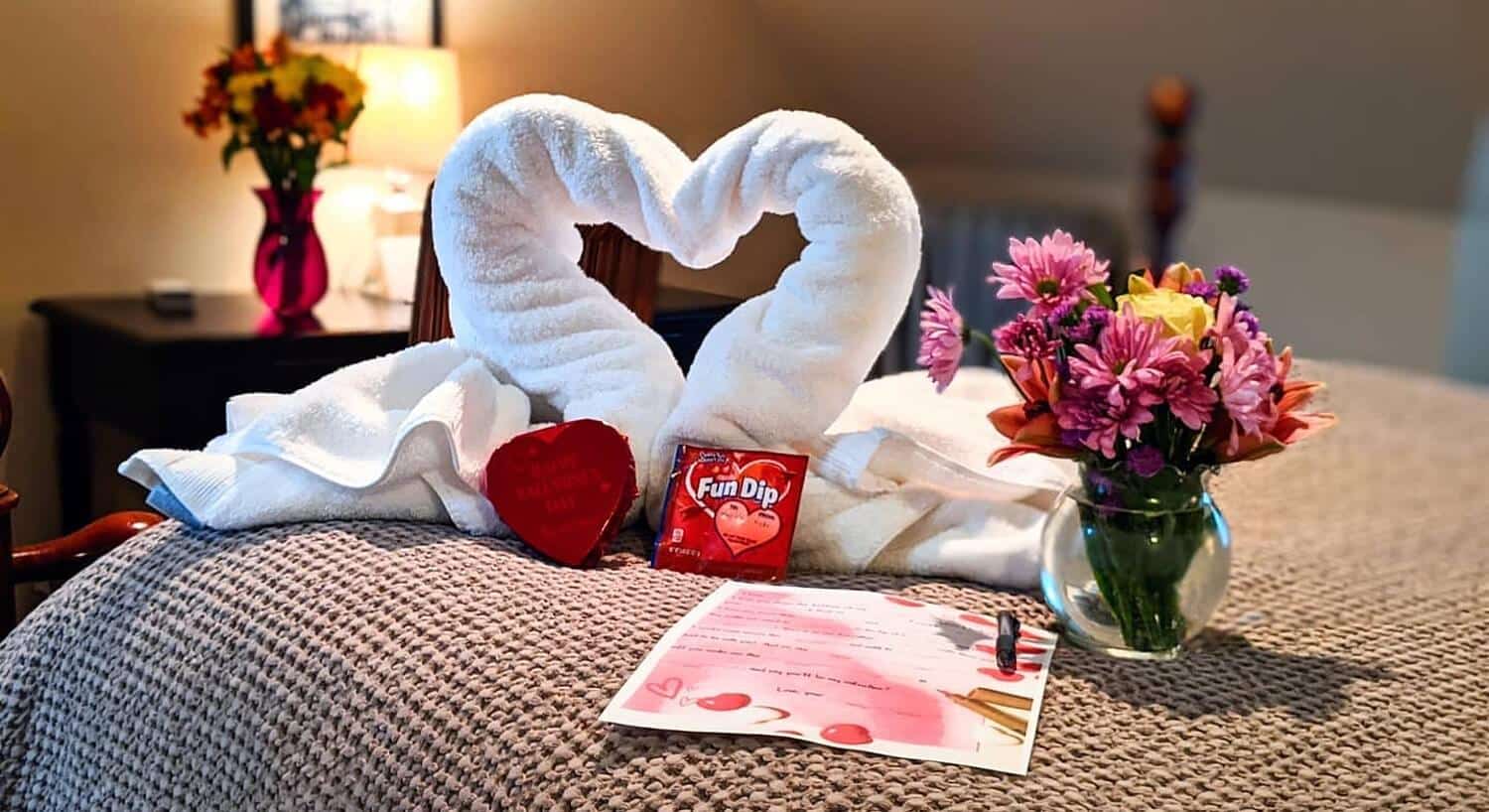 A white towel in the shape of a heart on a bed with vase of flowers, candy and a letter