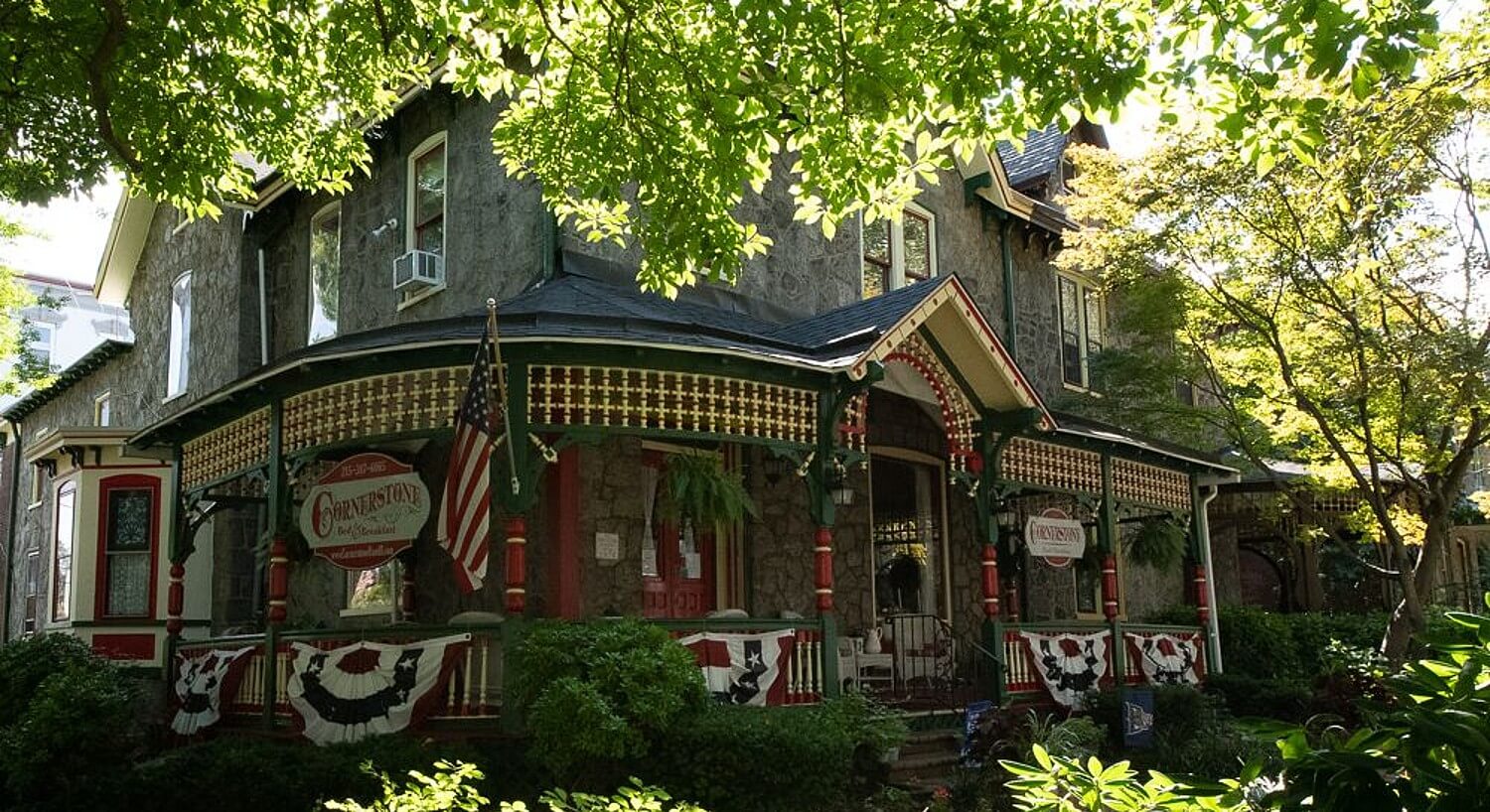 Front facade of a home with stone walls, large wrap around porch surrounded by lush landscaping and mature trees