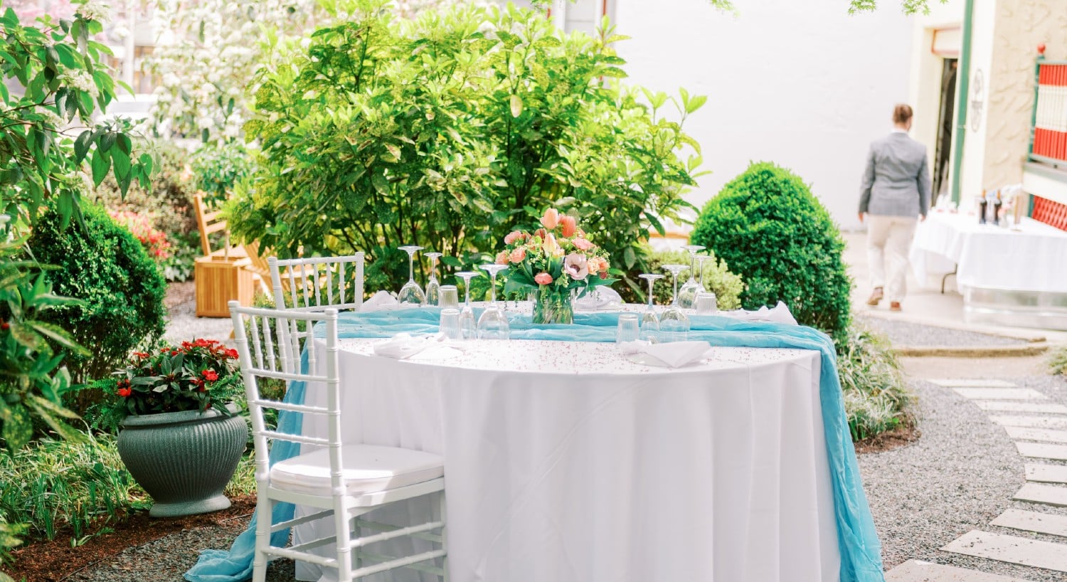 A round table with white linens, set for a wedding reception on an outdoor patio surrounded by lush greenery