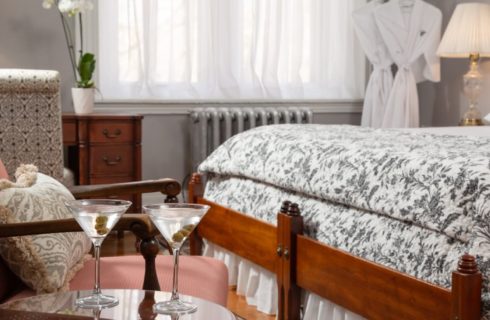 Salmon colored sitting chair by a table holding two martinis in a bedroom with king bed and hanging robes