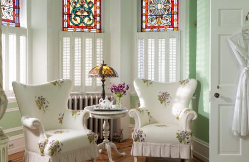 Two flowered wingback chairs under beautiful stained glass windows near open door with a hanging robe