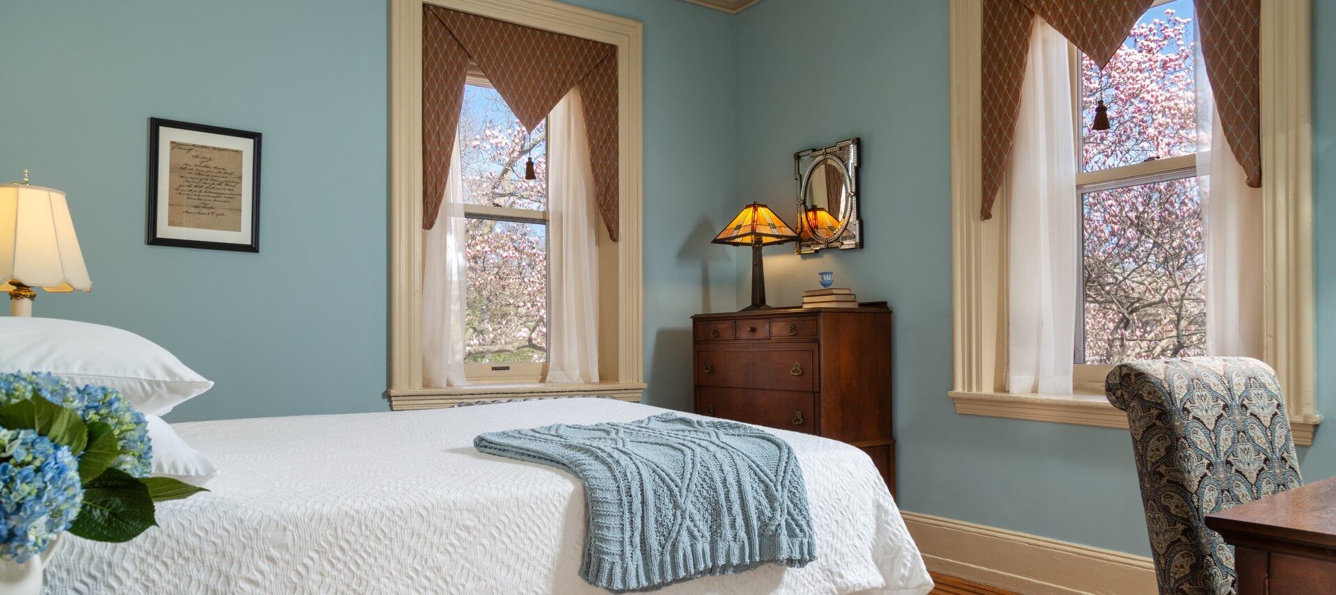 Guest room with white bed, two large windows, dresser and sitting desk with chair