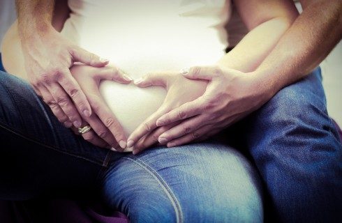 A pregnant woman in a white t-short making a heart shape with her hands on her stomach