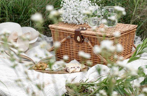 A brown picnic basket with a wooden plate of snacks on a white blanket in the grass