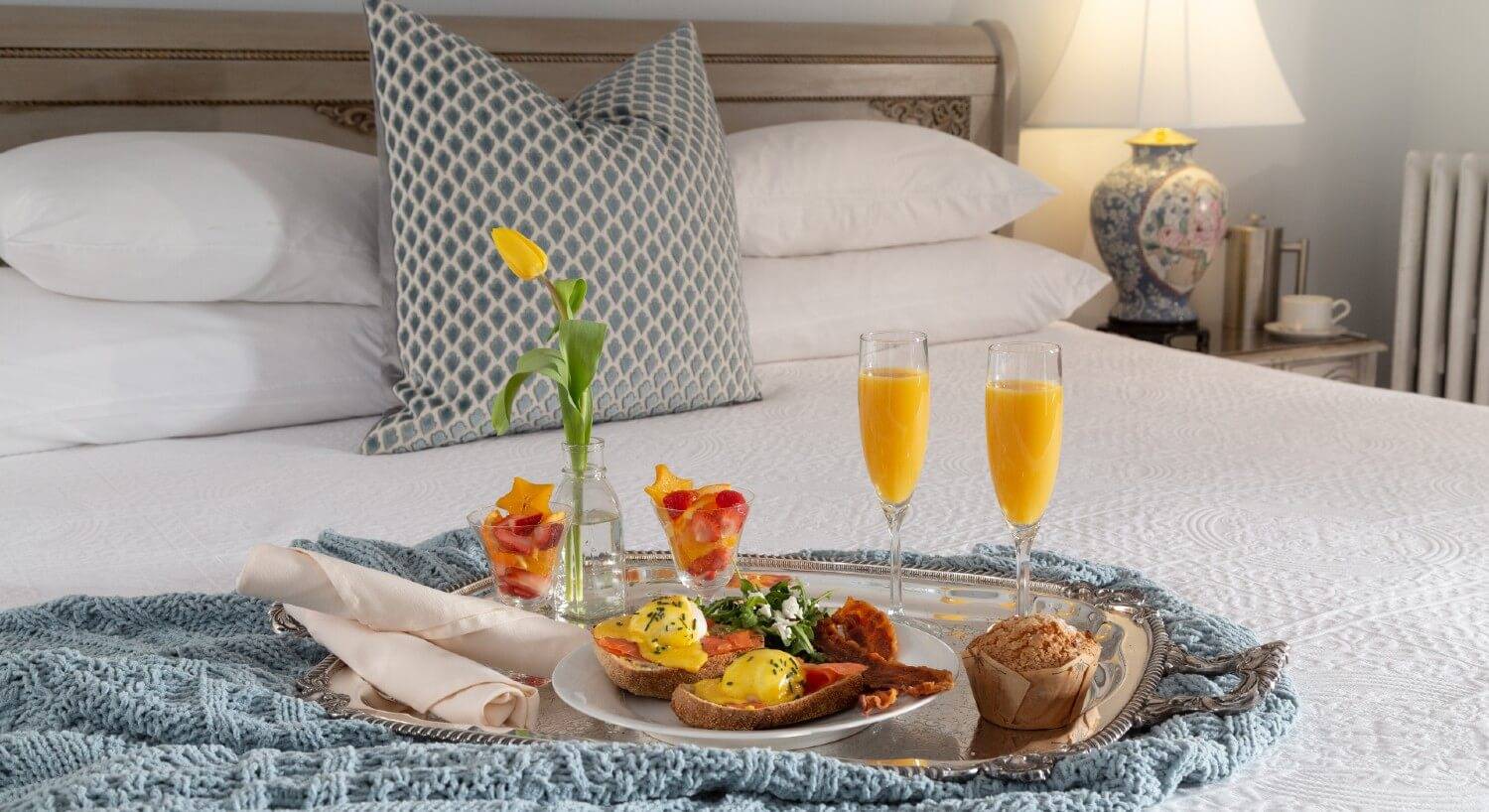 Bed with silver platter with plate of gourmet breakfast, fruit cups, muffin and two glasses of orange juice
