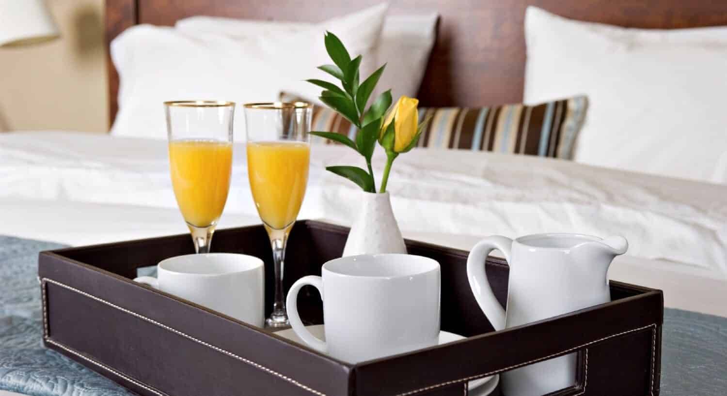 Brown leather tray on a bed holding two flutes of orange juice, two white coffee mugs and a creamer