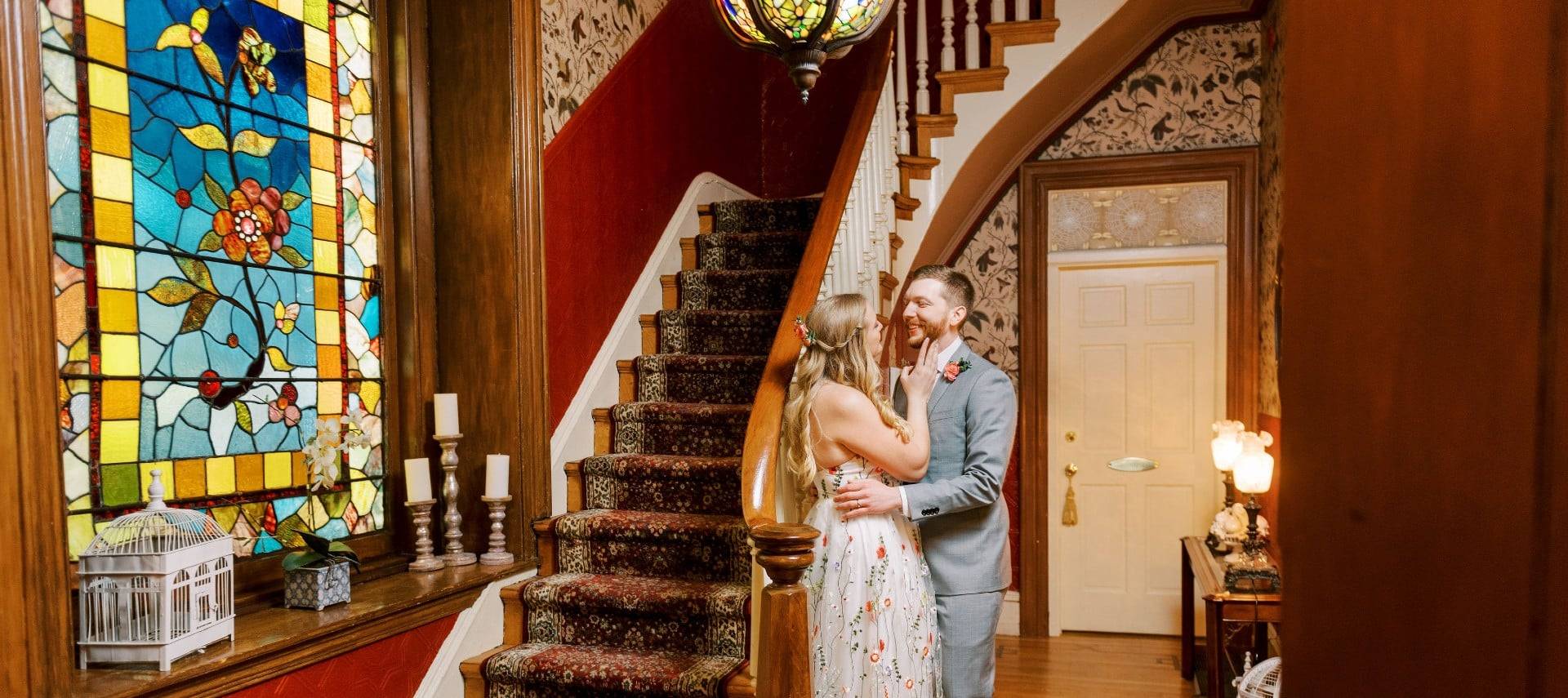 A bride and groom standing in a stunning front entry of a home with stained glass window and curved staircase